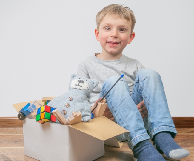 boy in jeans, tshirt and socks sitting next to a box of toys which include a teddy bear, a Rubix cube and a wooden train and tracks